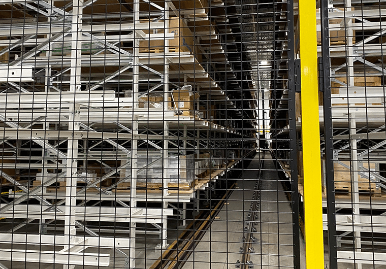 ASRS 3 Warehouse aisle with tall metal shelving, pallets stacked on racks, viewed through a metal mesh gate with a yellow post on the right edge